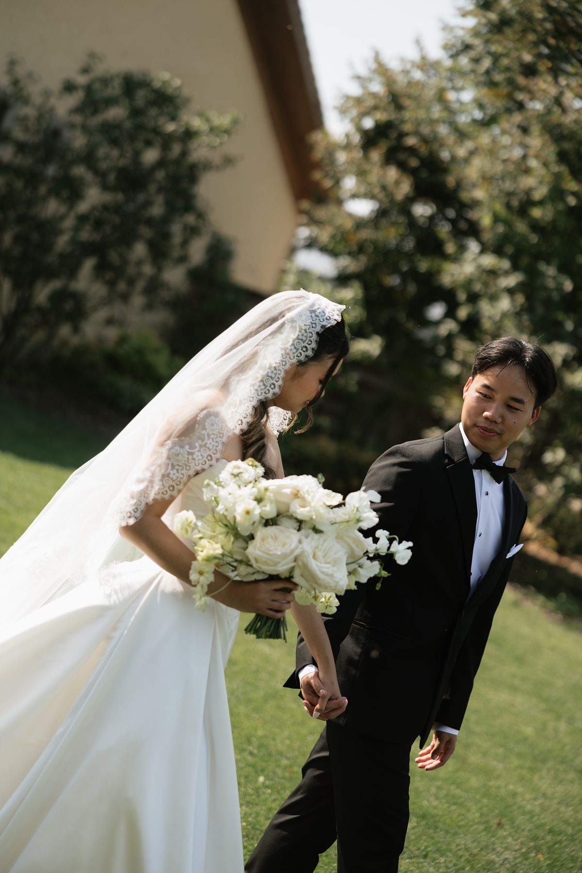 A bride in a white lace gown holding a white bouquet walks with a groom in a black tuxedo on a green lawn.