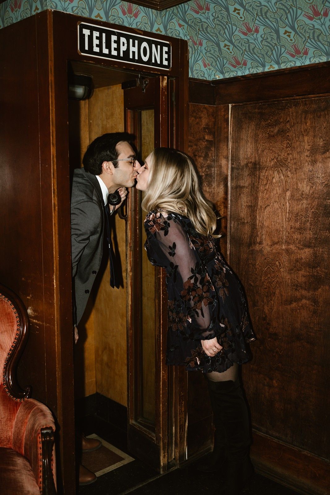 A couple shares a romantic moment by a vintage telephone booth in a room with patterned wallpaper and wood paneling.