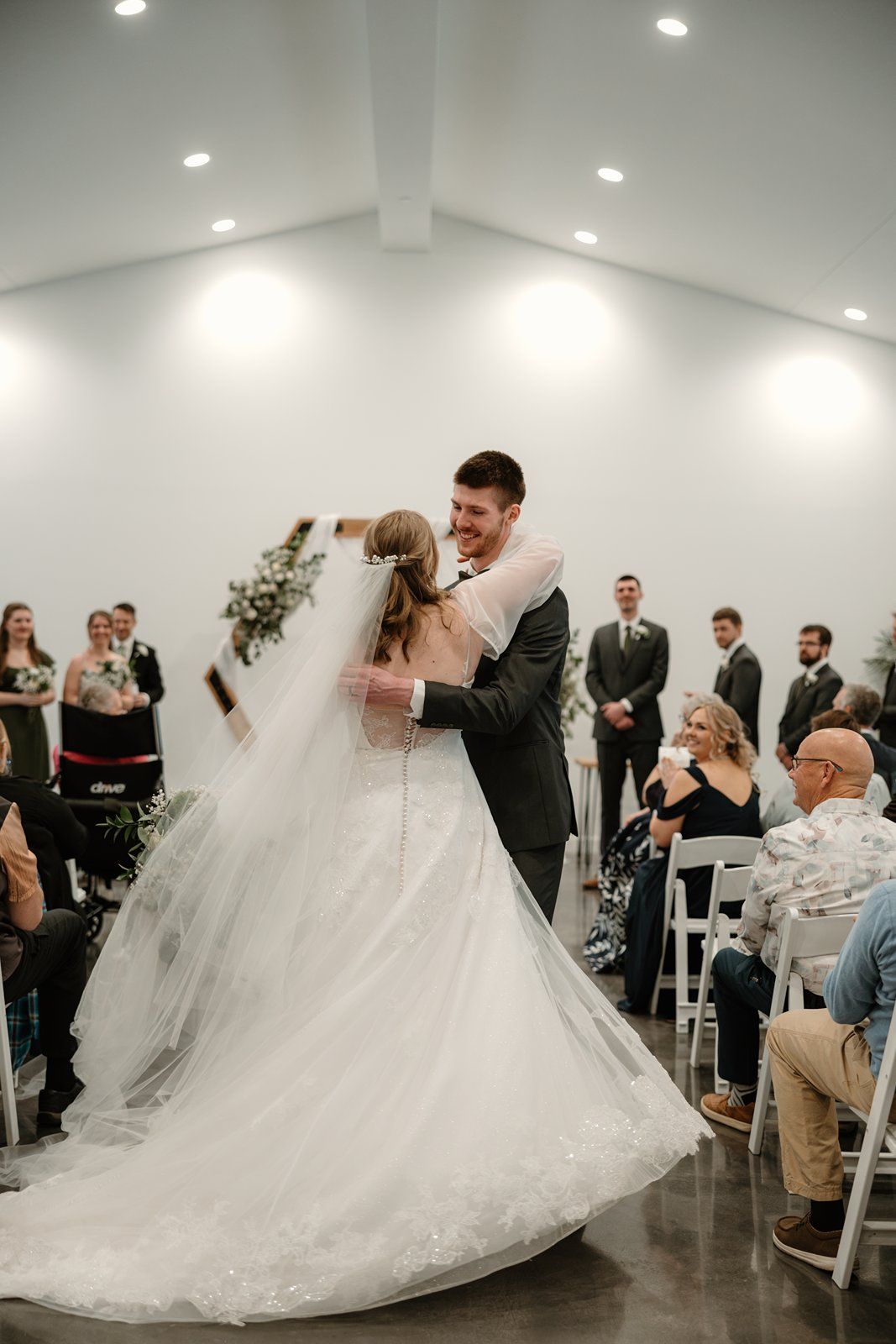 A bride and groom share their first dance at a modern indoor wedding venue with guests watching from white chairs.