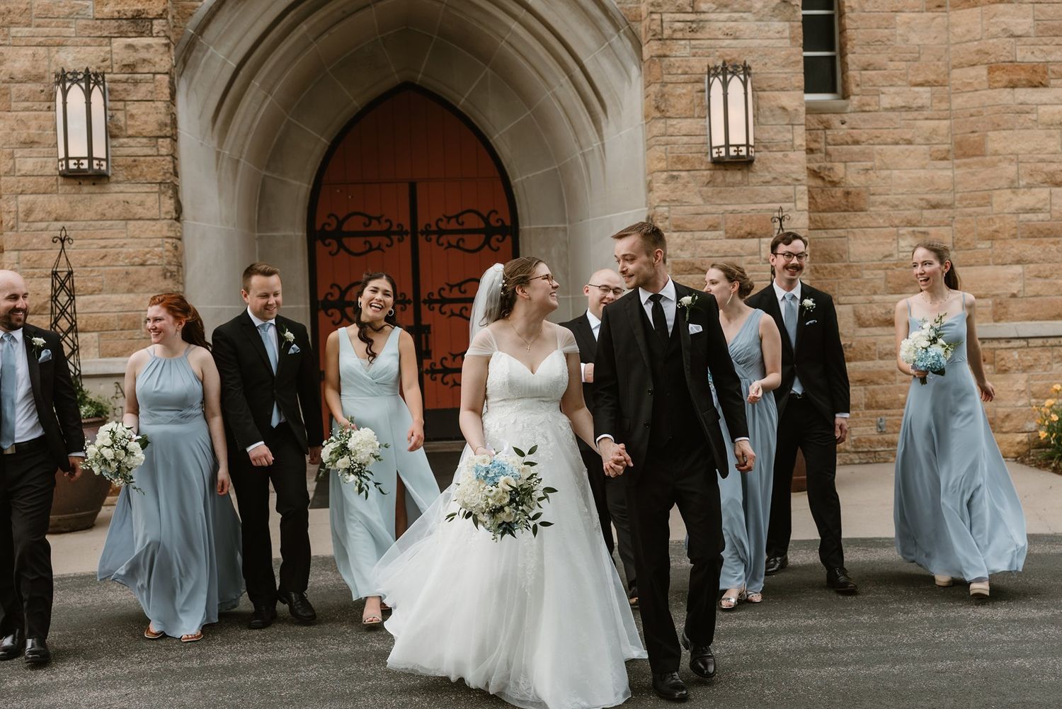 Wedding party in formal attire walks together outside stone church with arched doorway in background.