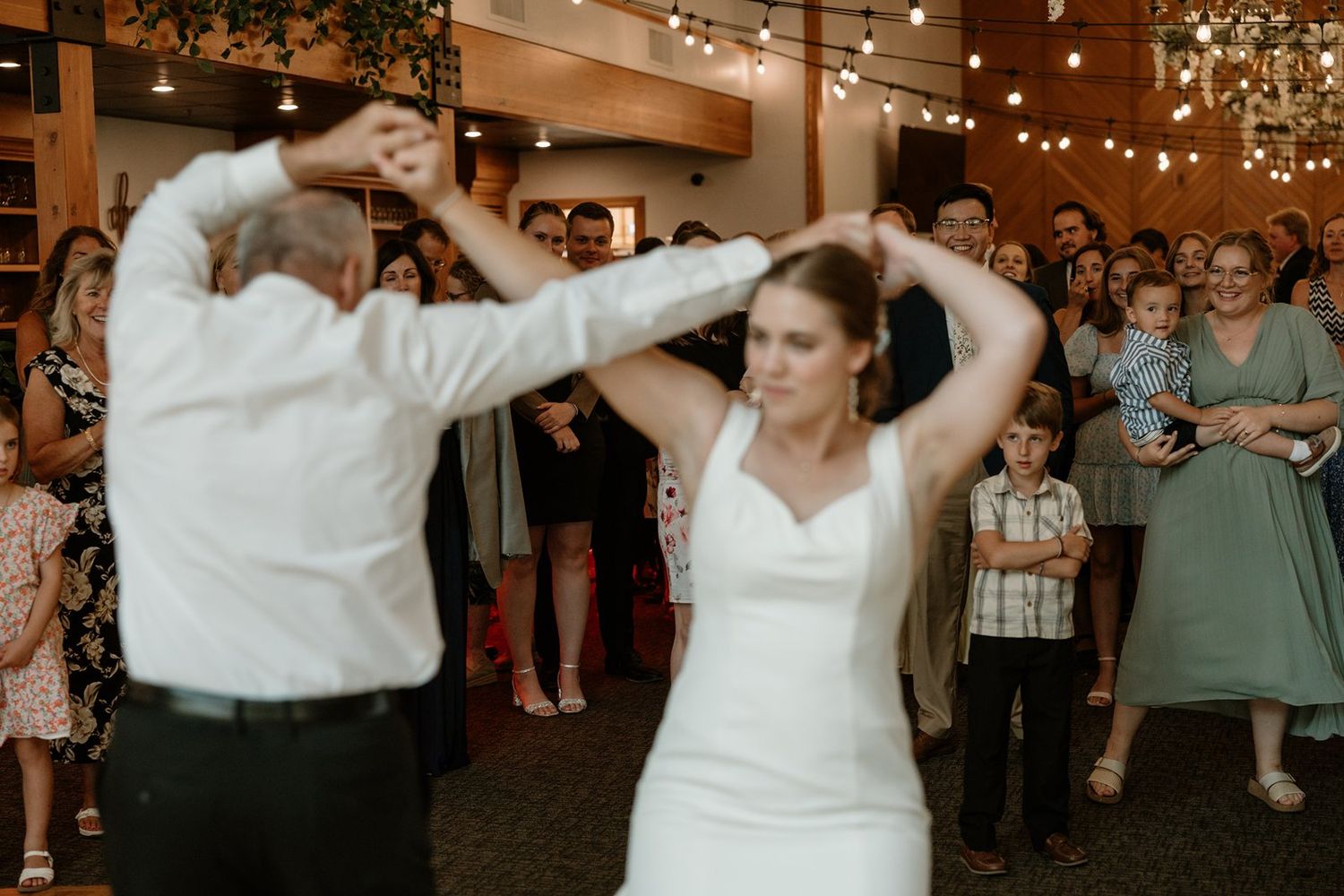 Wedding guests dancing and celebrating on a dance floor decorated with string lights overhead at a reception.