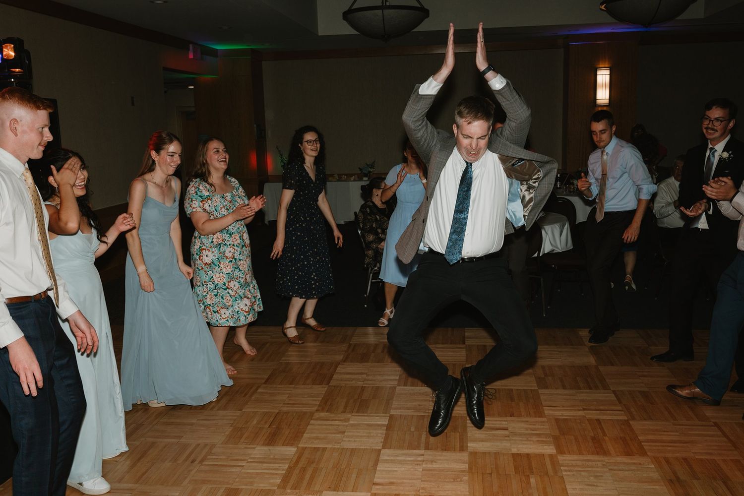 A person leaps high while dancing on a parquet floor at an indoor celebration with others watching.