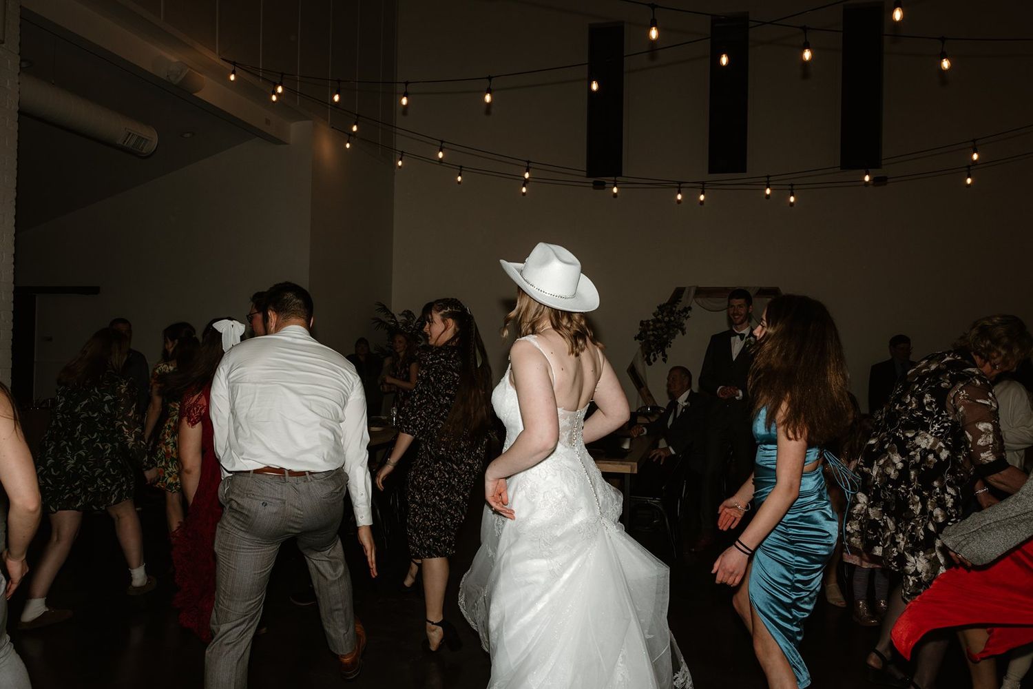 Wedding guests dancing and celebrating on a dimly lit dance floor at an indoor reception venue.