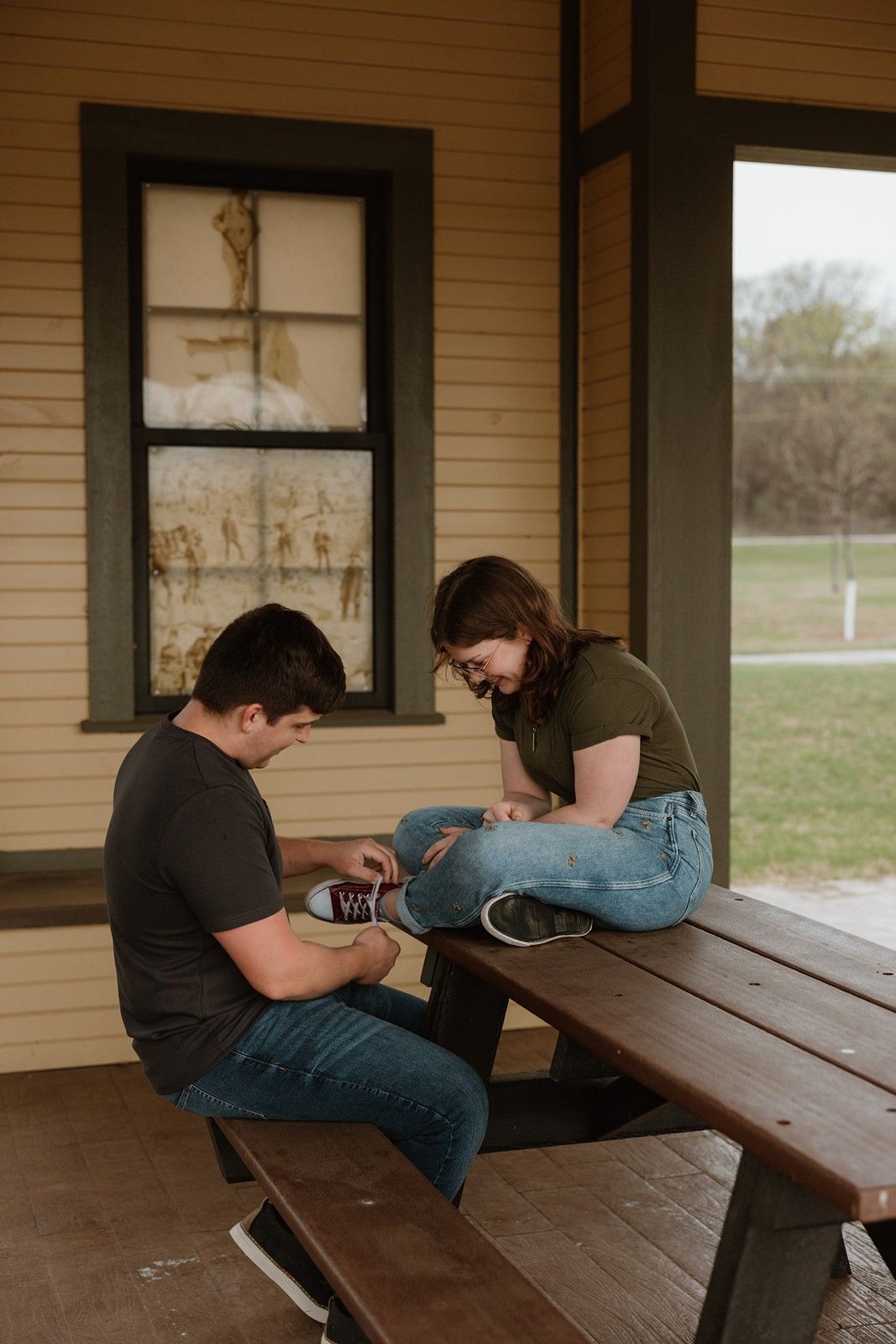 Two people sit together on a rustic wooden picnic table on a covered porch, looking down at something between them.