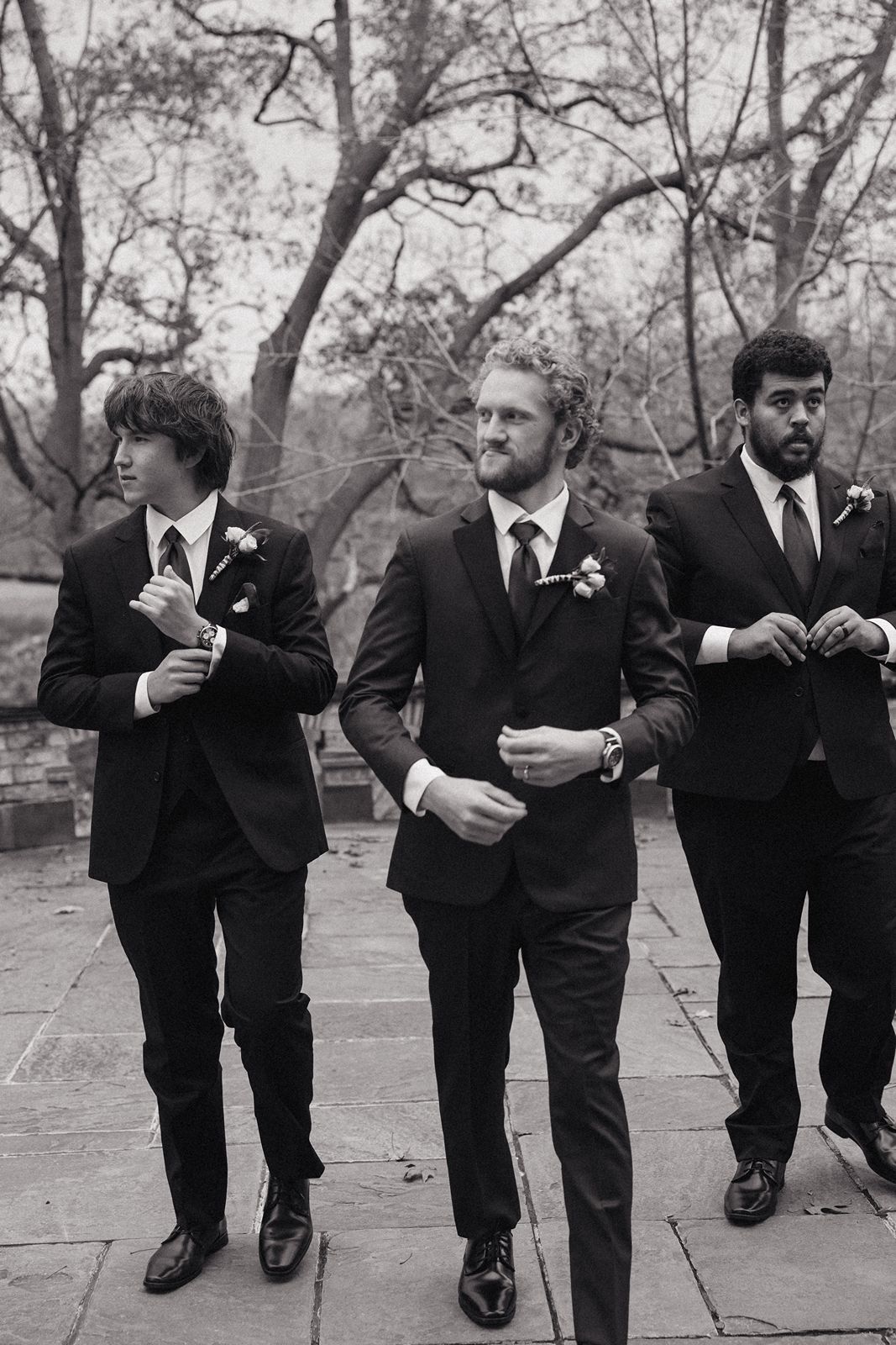 Three groomsmen in black suits walk together outdoors during wedding festivities.