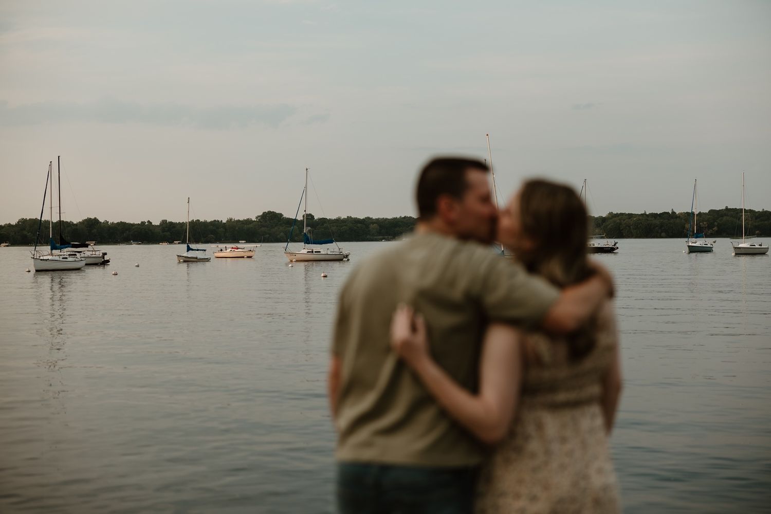 Couple sharing a romantic kiss by a serene lake with sailboats at dusk.