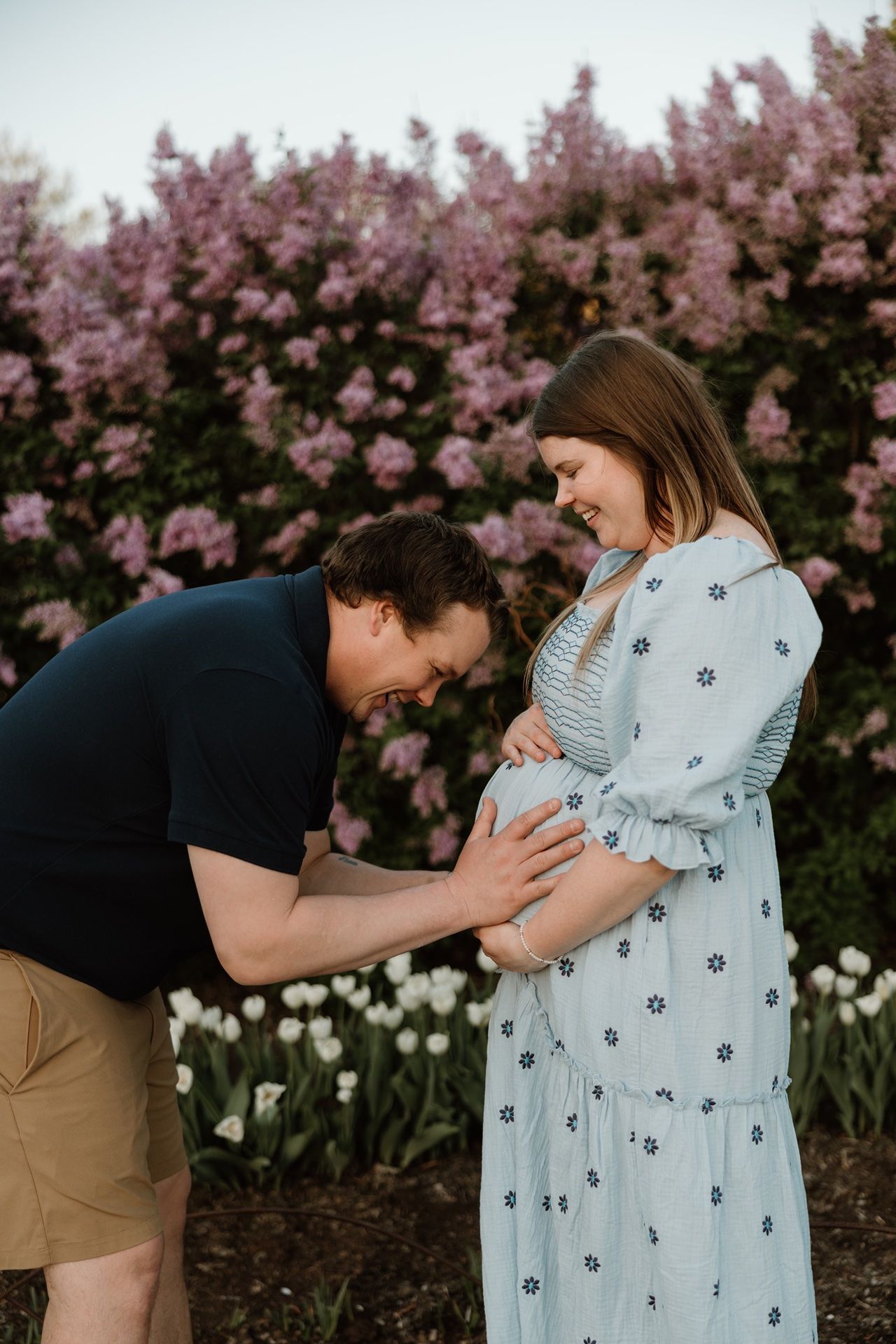 Expectant couple sharing a tender moment outdoors, man leaning to kiss pregnant belly, pink flowering bushes and white tulips behind.