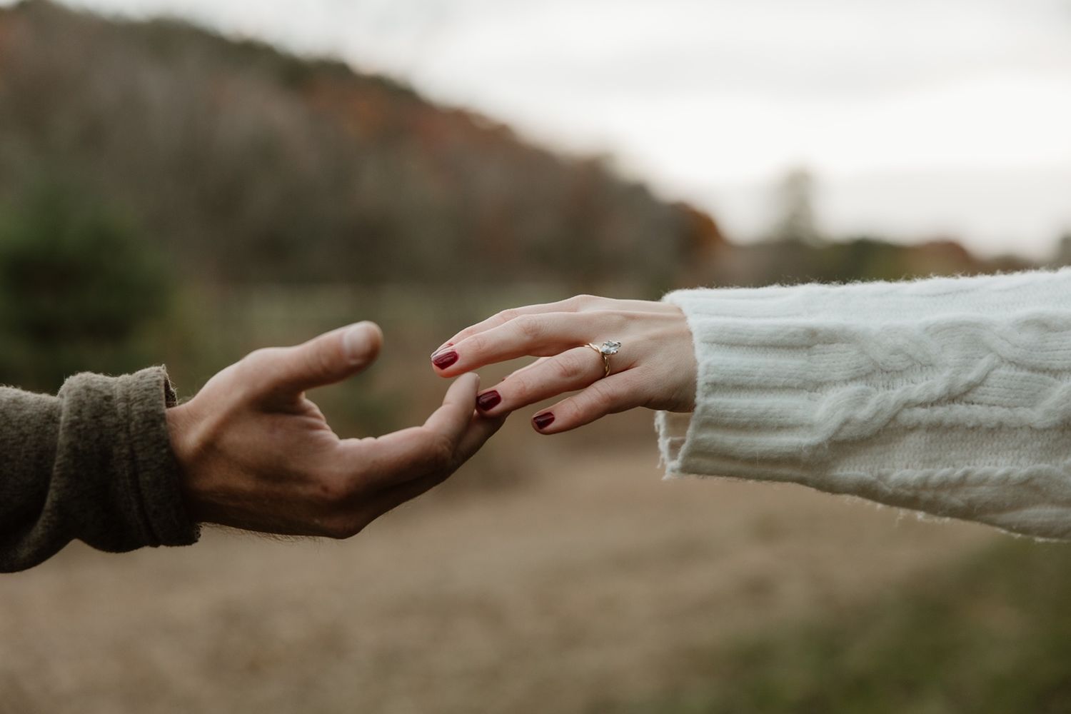 Two hands reaching toward each other outdoors, one wearing a white sweater with a ring, set against a blurred natural background.