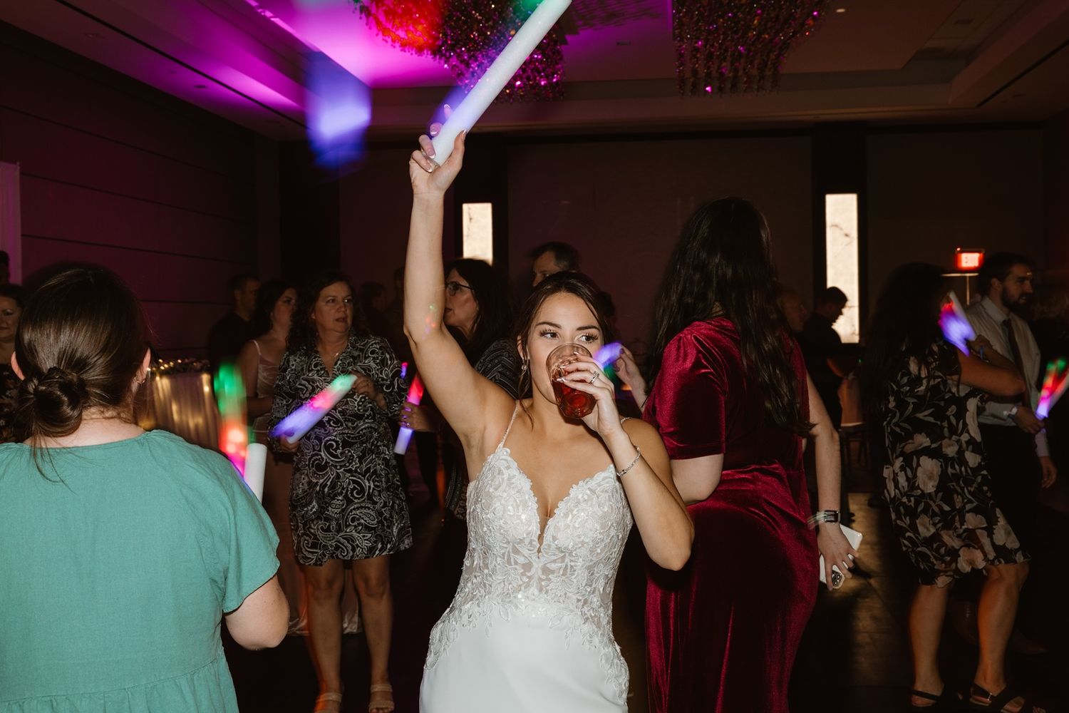 Bride in white gown dancing joyfully at wedding reception, holding glow stick above head among guests.