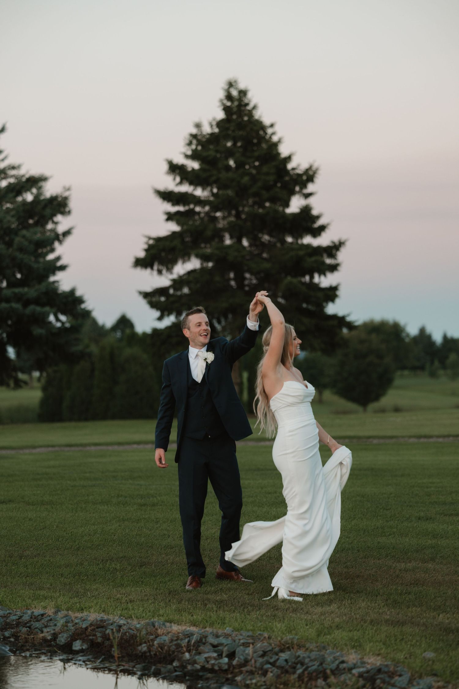 A couple in wedding attire dances playfully together on a grassy field at sunset with evergreen trees in the background.