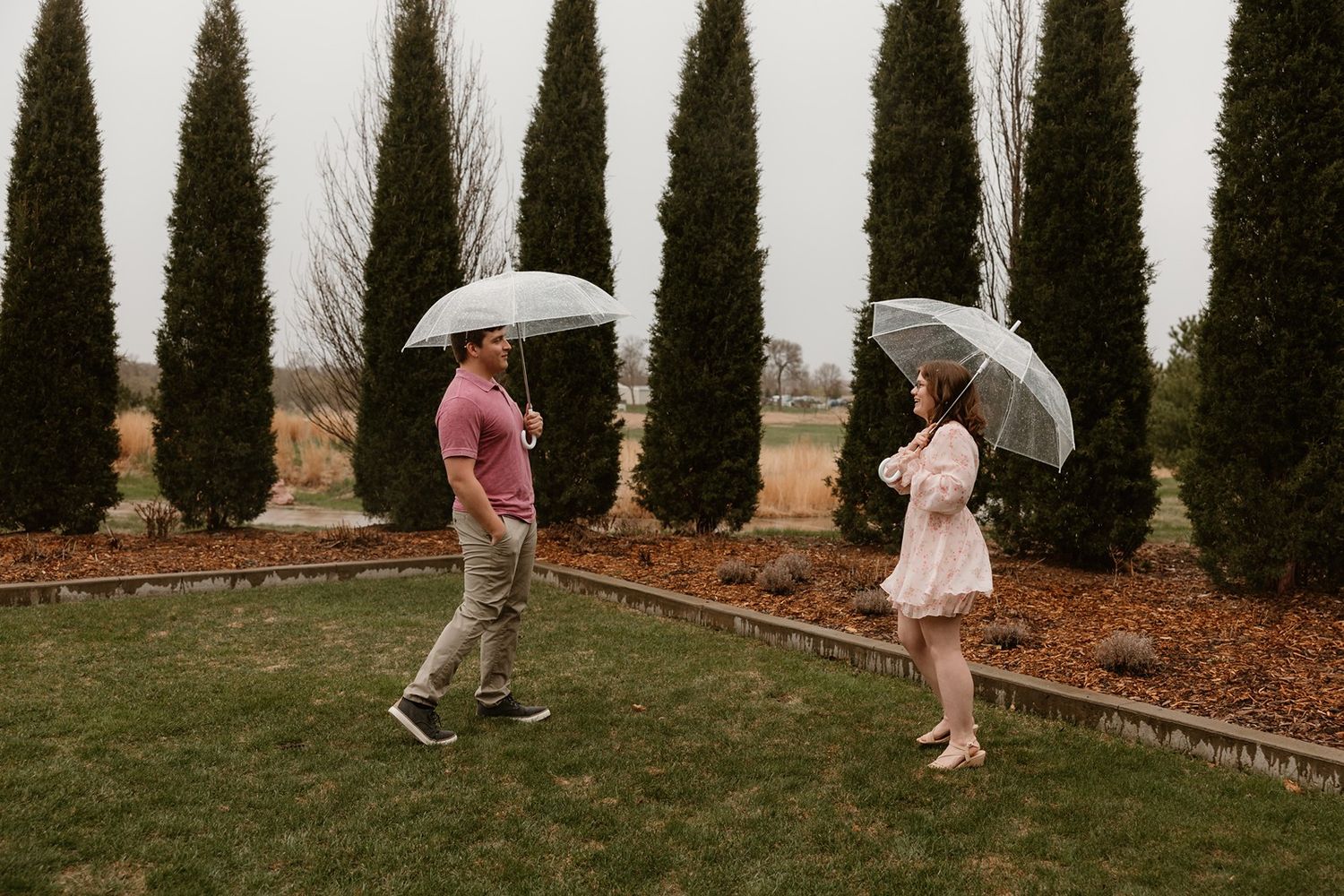 A row of evergreen trees creates a backdrop for multiple people dressed in formal attire walking on grass.