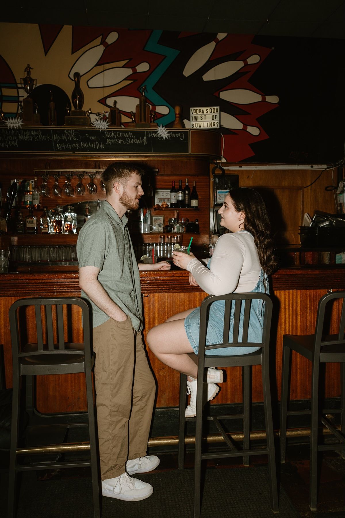 A couple shares playful moments at a dimly lit bar counter with wooden decor and stools.