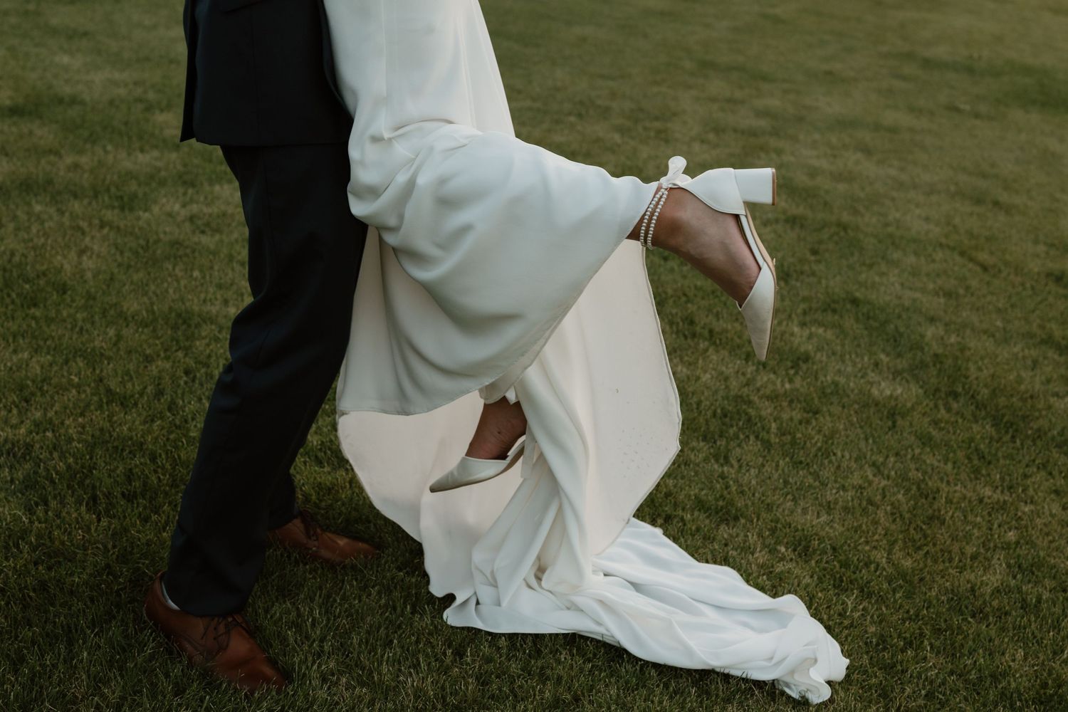 A couple in white wedding attire walks together on a lush green lawn during their outdoor celebration.