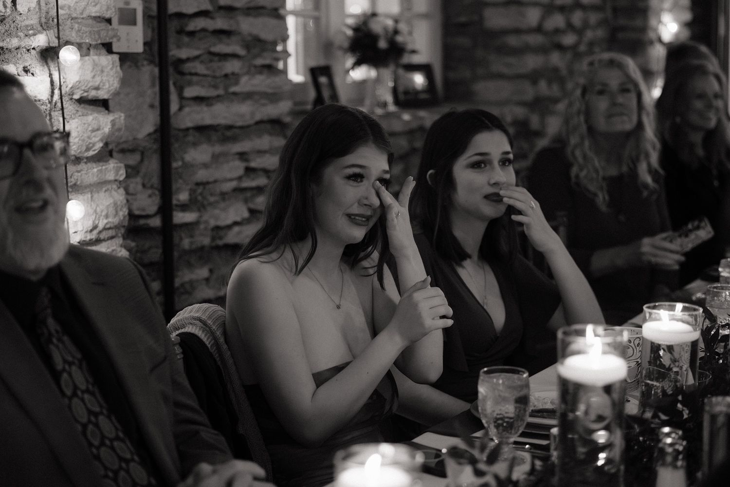 Black and white photo of women tearing up during emotional speeches at a candlelit reception.