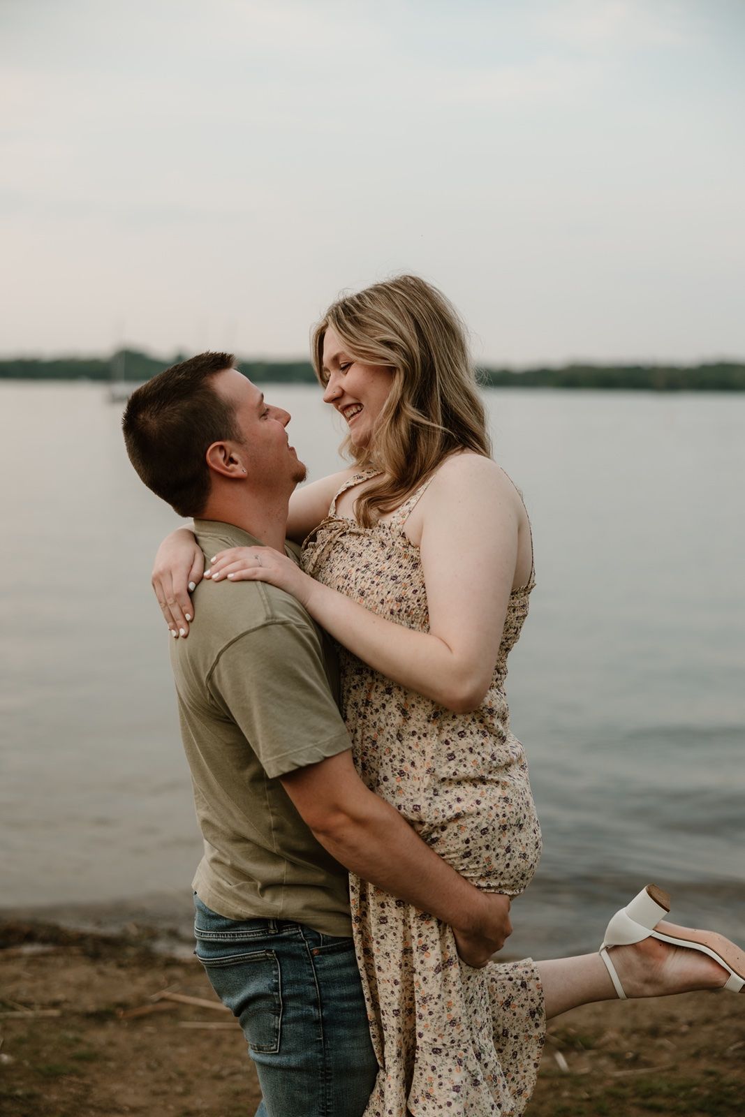 A couple shares a joyful embrace by a lakeside, with the woman laughing as her partner lifts her gently.
