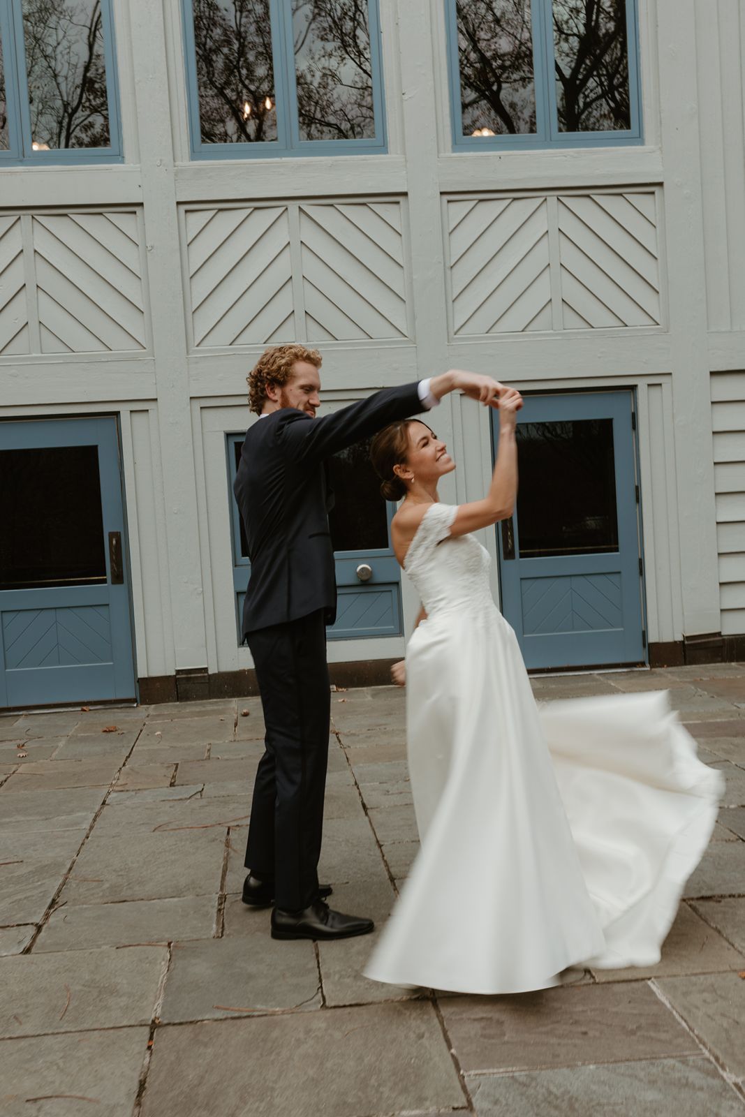 Couple performs a romantic dance spin on stone patio in front of a geometric white building.