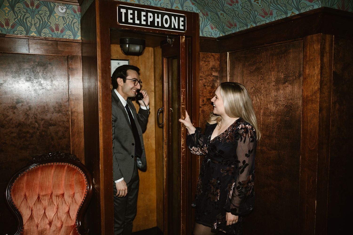 Two people in stylish dark attire conversing near a vintage wooden telephone booth with orange velvet chair nearby.