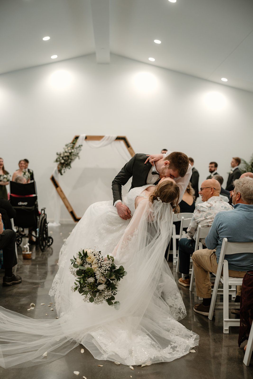 A romantic wedding dip kiss captured during a ceremony in a modern white venue with floral decorations and seated guests.
