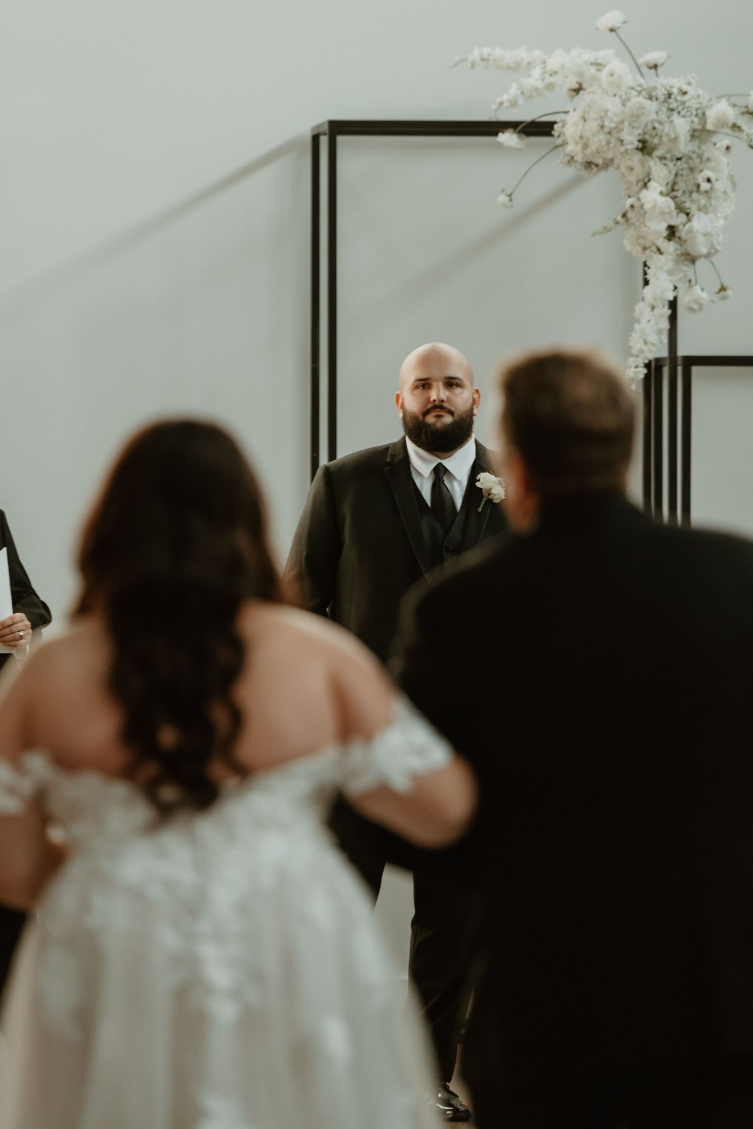 A tender moment captured during a wedding ceremony with white floral decor and minimal modern black framed backdrop.