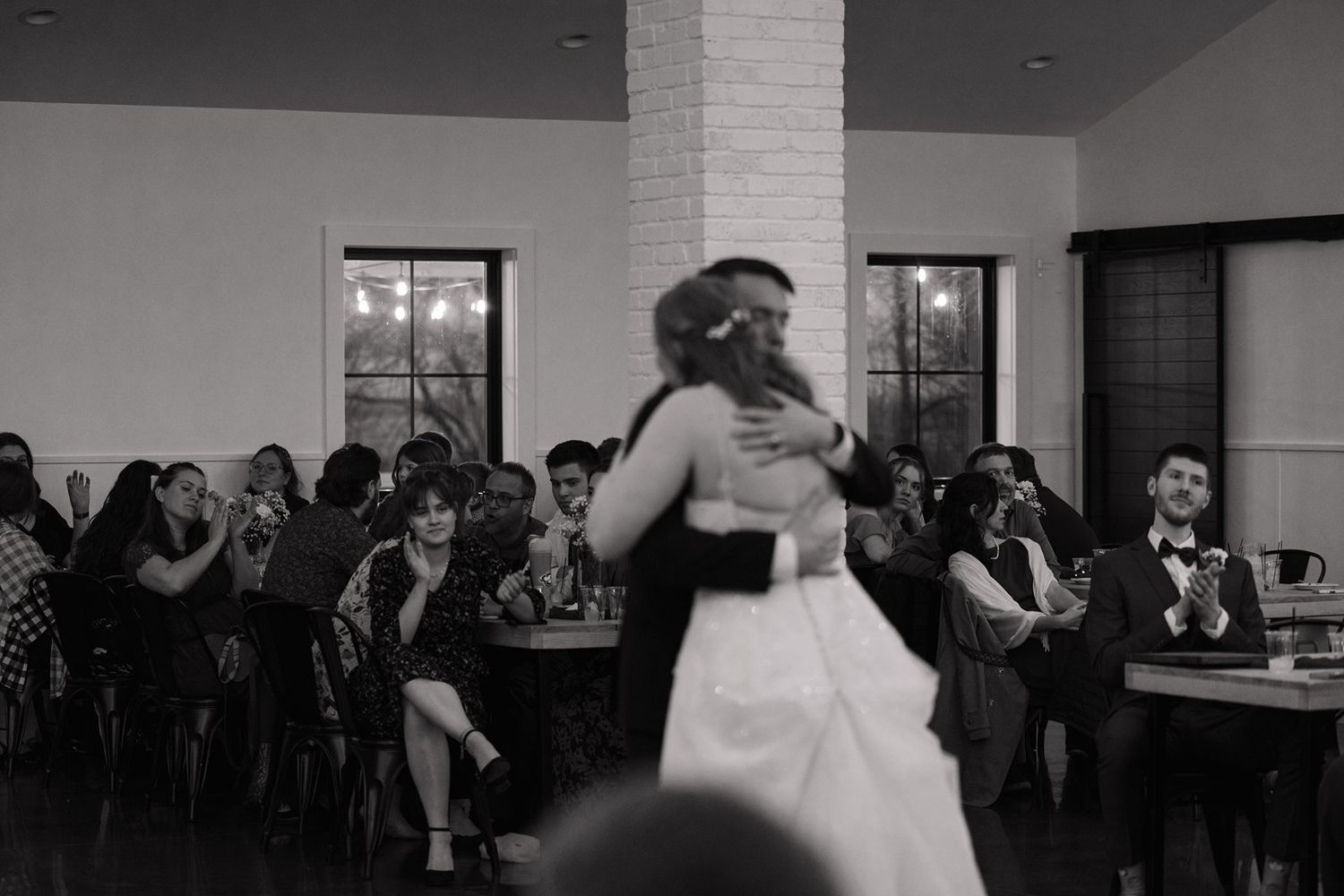 A black and white photo of a couple sharing their first dance at a wedding reception with guests watching.