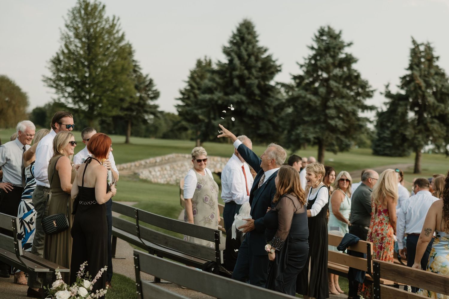 Wedding guests gather outdoors under tall evergreen trees during a summer celebration ceremony.