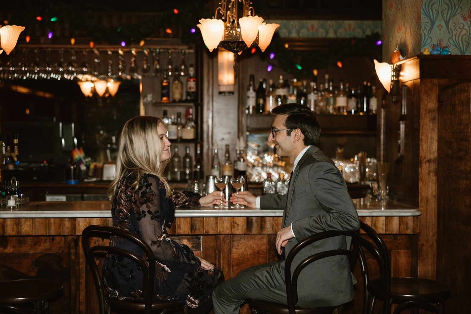 A couple shares drinks at a vintage bar with ornate lighting fixtures and classic decor.