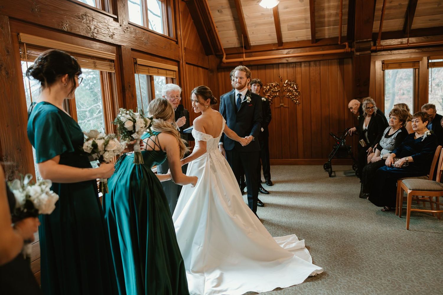 A wedding ceremony takes place in a rustic wooden chapel with bridesmaids in teal and black dresses.