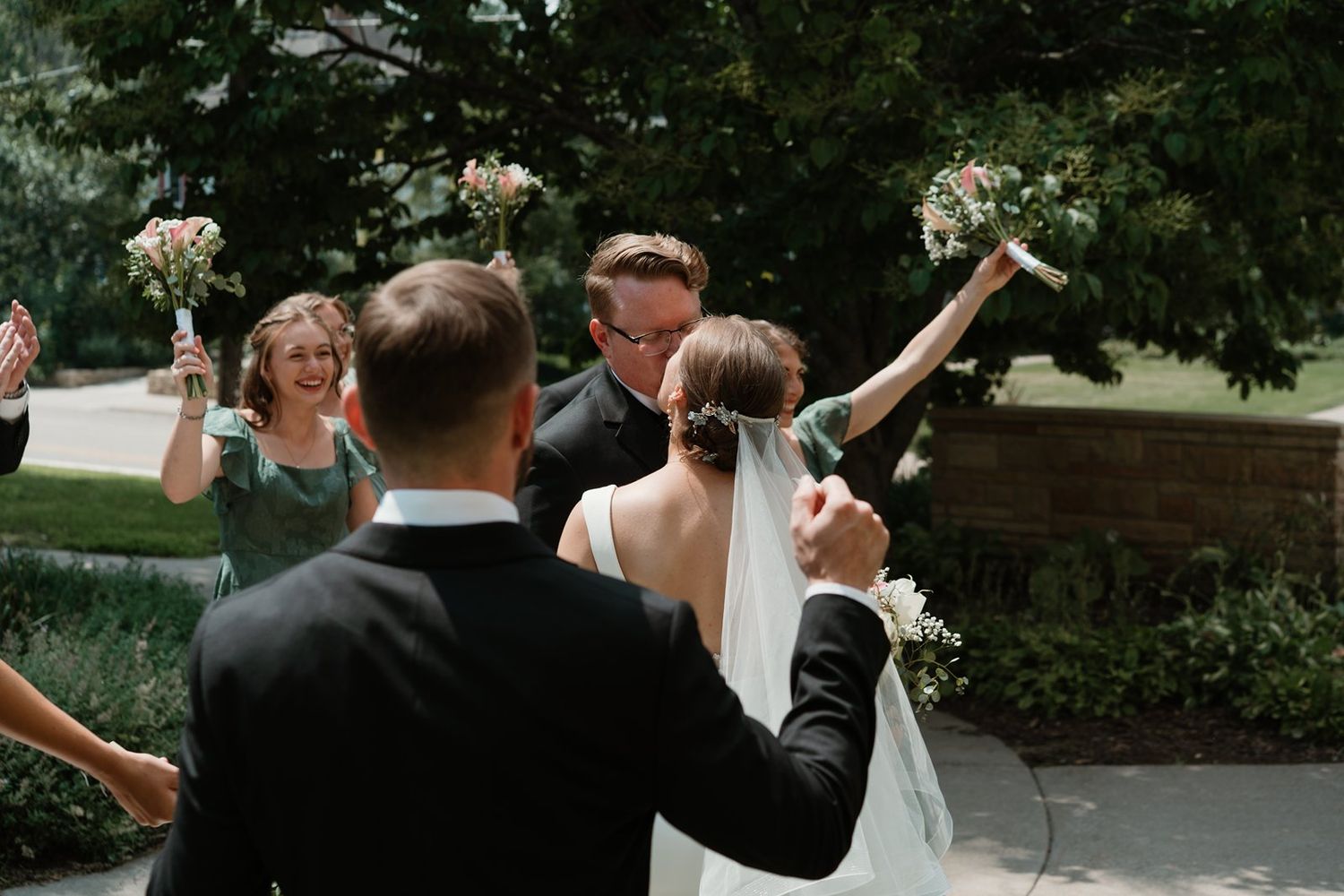 Wedding party celebrates joyfully outside with raised arms and bouquets against green foliage background.