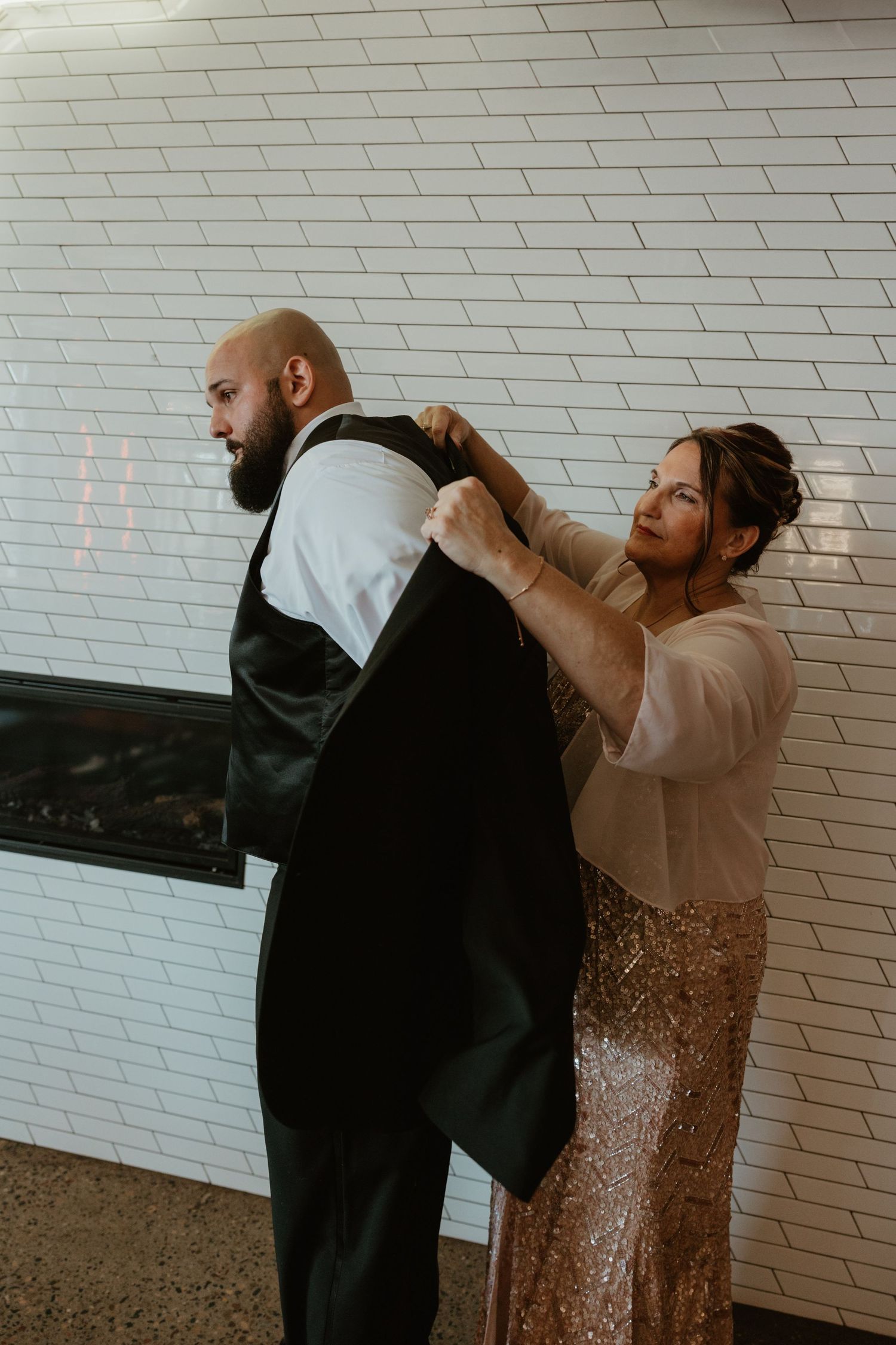 A couple getting ready for a formal event against a white brick wall, with her adjusting his black suit jacket.