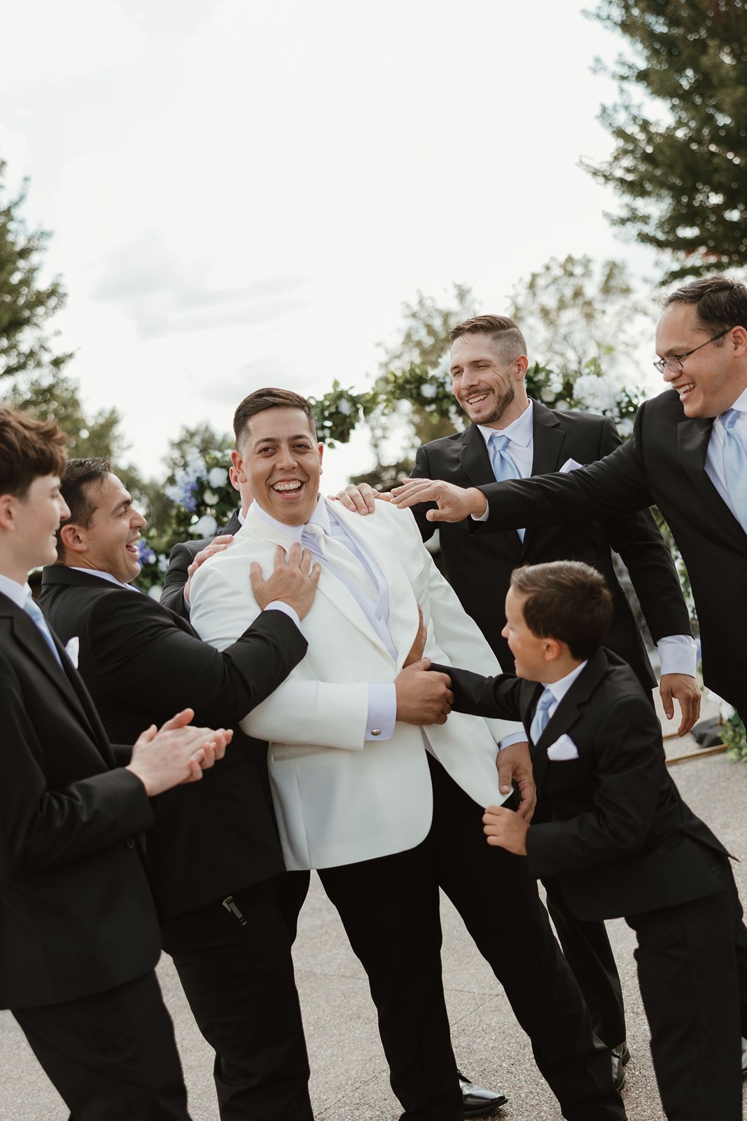 Groomsmen in black suits share a lighthearted moment with the groom in white.