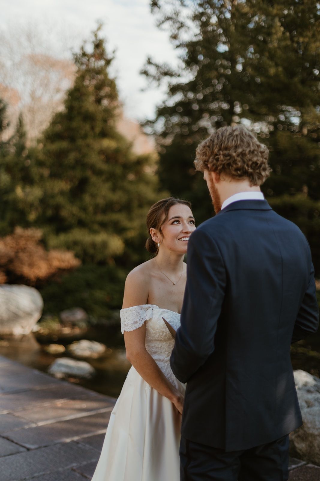 Bride in off-shoulder lace gown smiles at groom in navy suit during outdoor wedding ceremony near a serene garden pond.