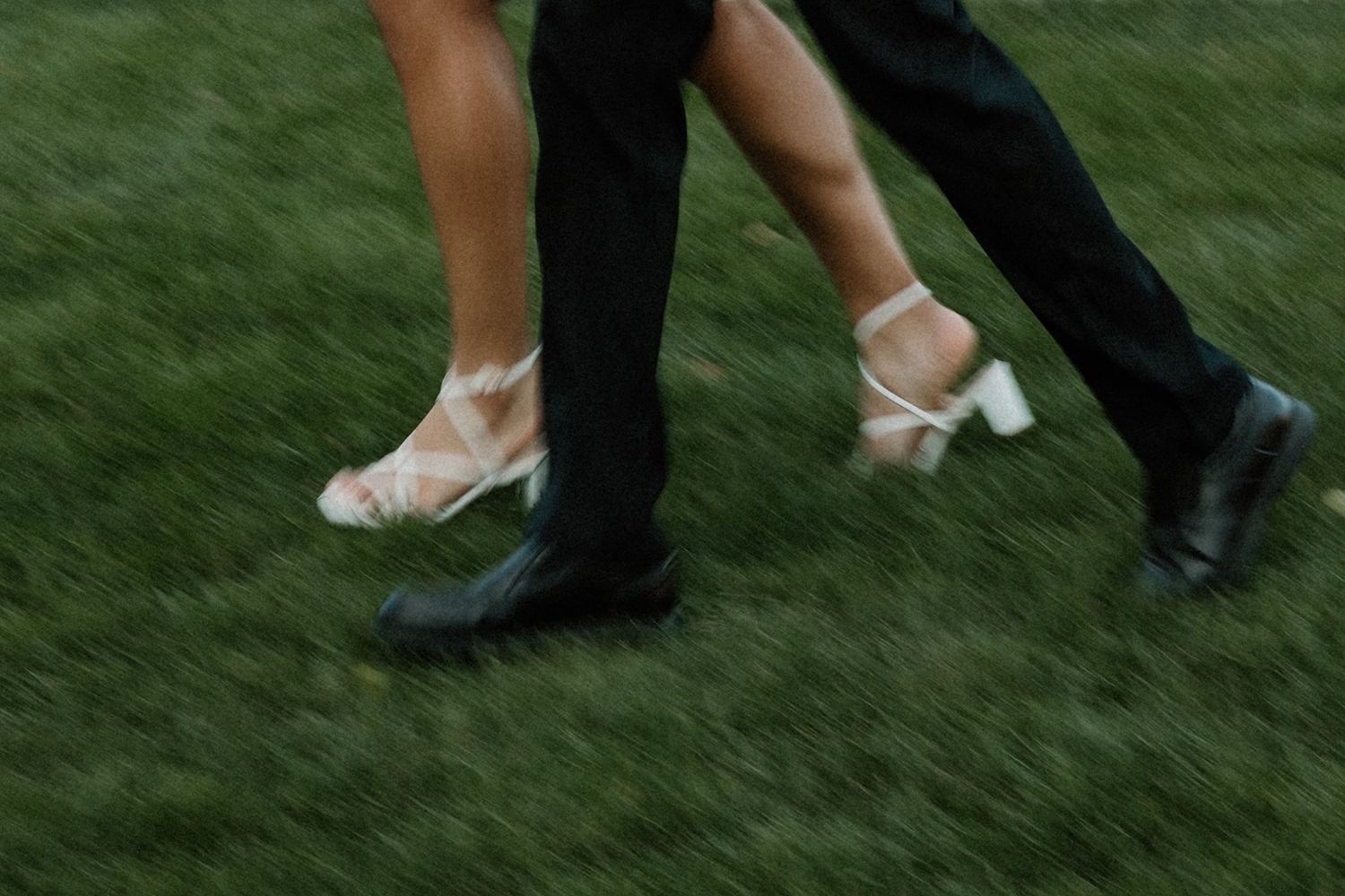 Close-up of a couple's feet dancing on grass, woman in white heeled sandals and man in black dress shoes.