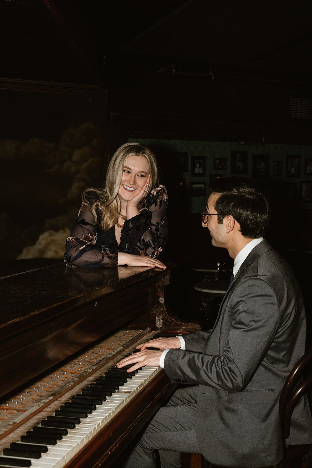 Elegant couple shares a moment at a piano in a dimly lit vintage setting.