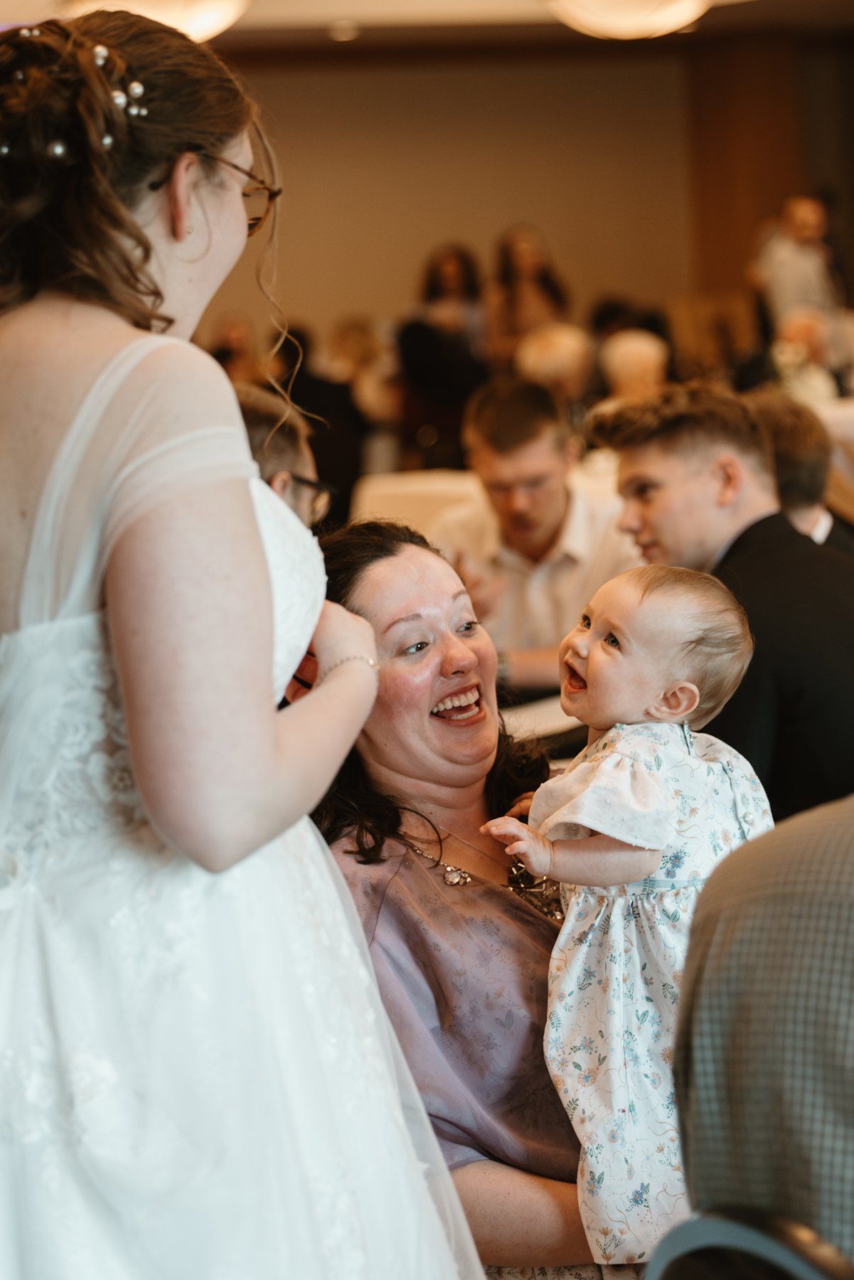 A joyful candid moment at a wedding reception shows guests sharing laughter while holding a baby in formal attire.