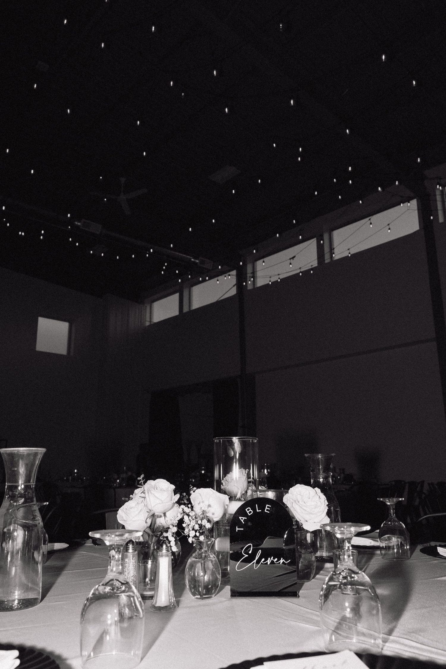 Black and white wedding reception table setup with white roses in glass vases under a starlit ceiling at night.
