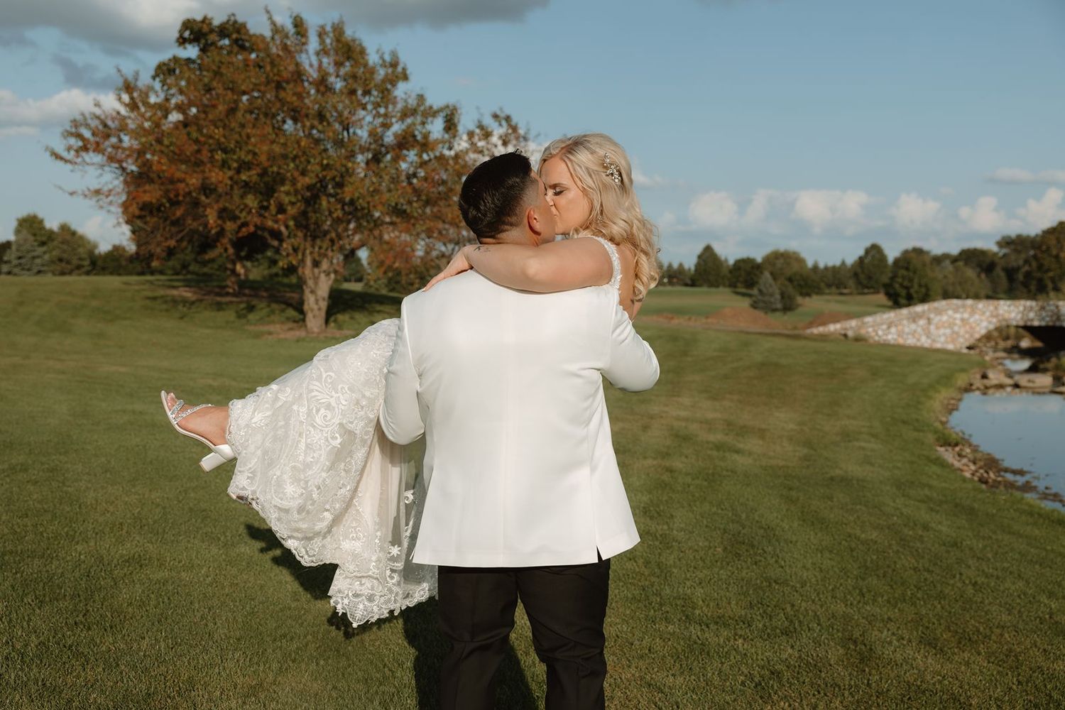 A groom in white jacket carries his bride across a scenic golf course during sunset.