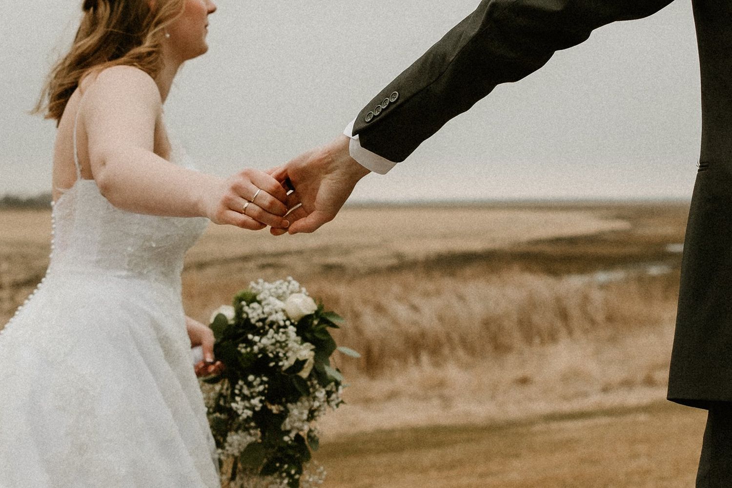 A couple holds hands during their wedding ceremony in a rustic outdoor setting with a spring farm landscape in the background.