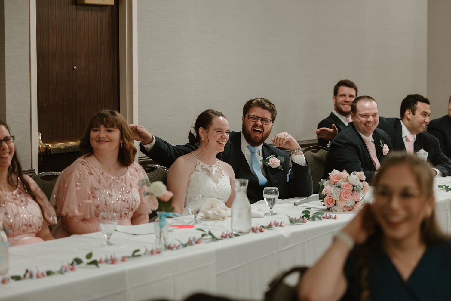 Wedding reception guests share laughter at the head table decorated with pink floral centerpieces.