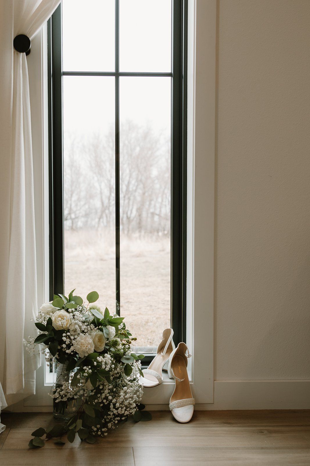 White bridal shoes and delicate floral bouquet arranged near bright windows in a minimalist room interior.