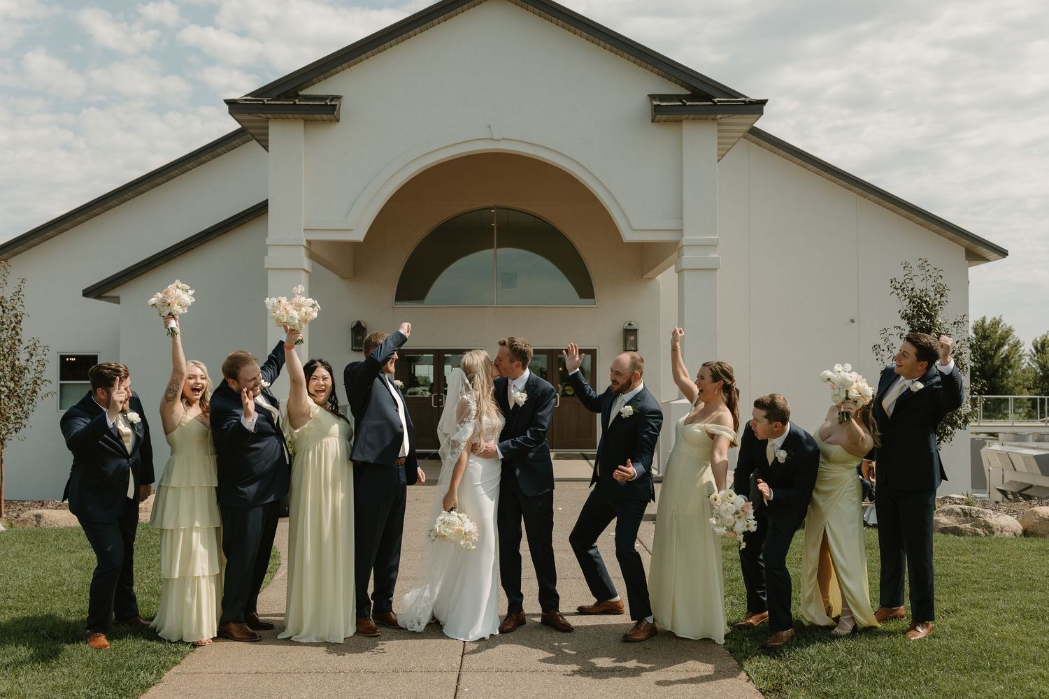 Wedding party members celebrate with raised arms in front of a white church building on a sunny day.