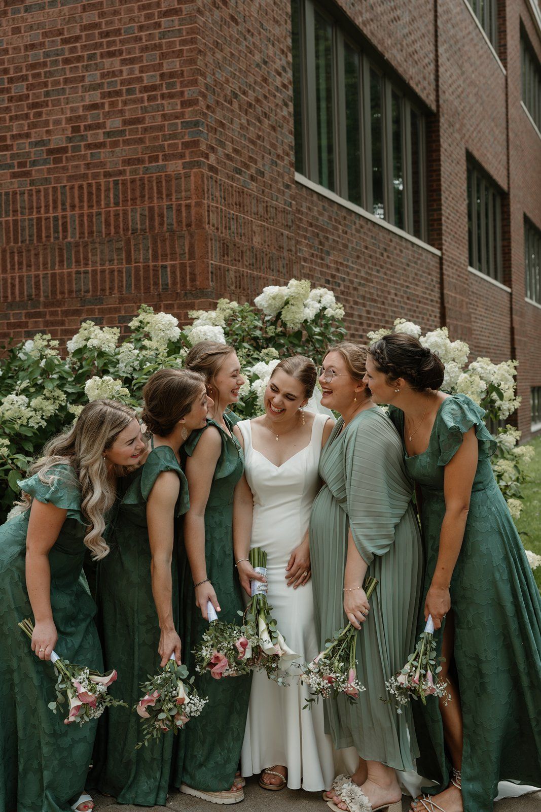 A wedding party wearing emerald green bridesmaid dresses and white bridal gown share candid laughter against brick wall.