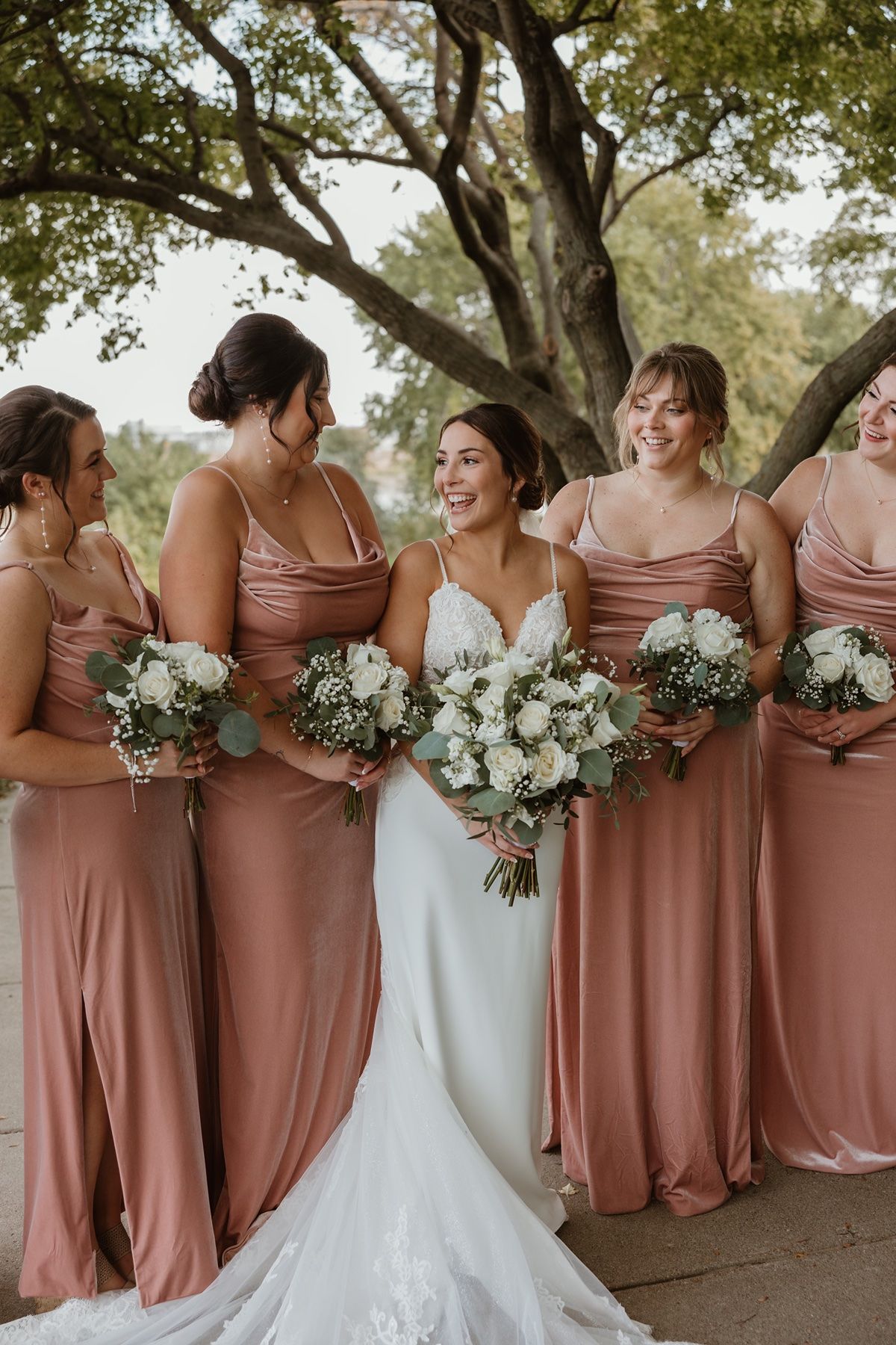 Bride in white lace gown smiles with bridesmaids in dusty rose satin dresses holding white floral bouquets outdoors.