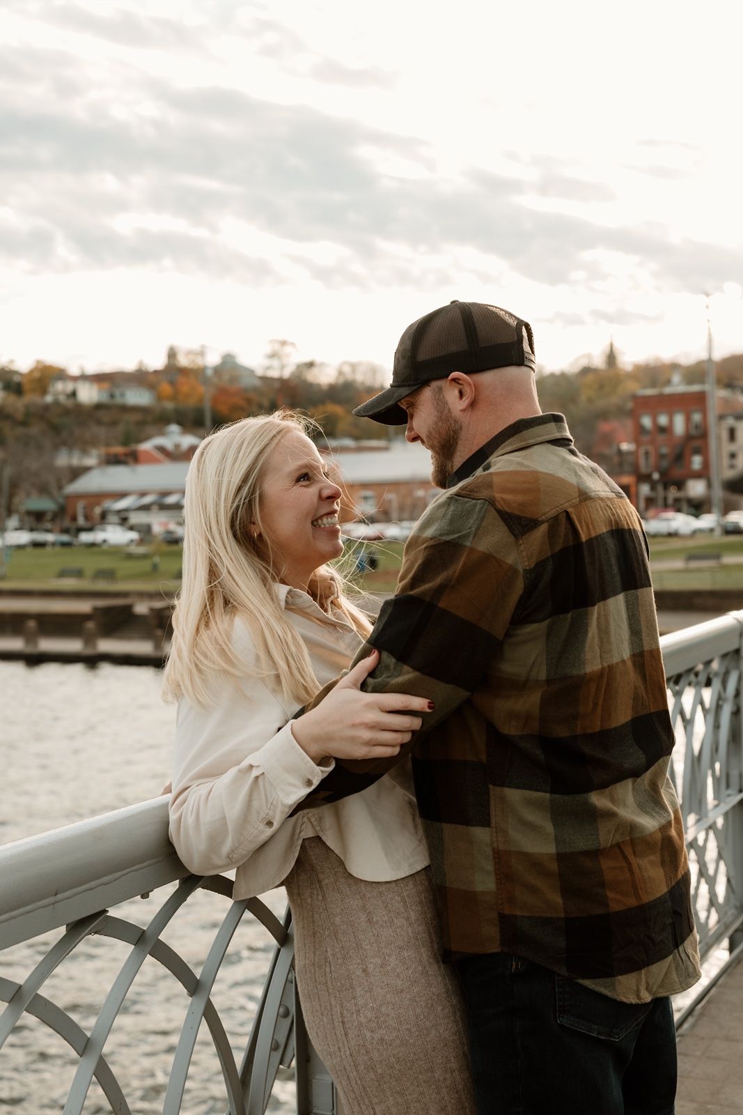 A couple shares a joyful moment on a bridge by the water, the woman in white and man in plaid embracing at golden hour.