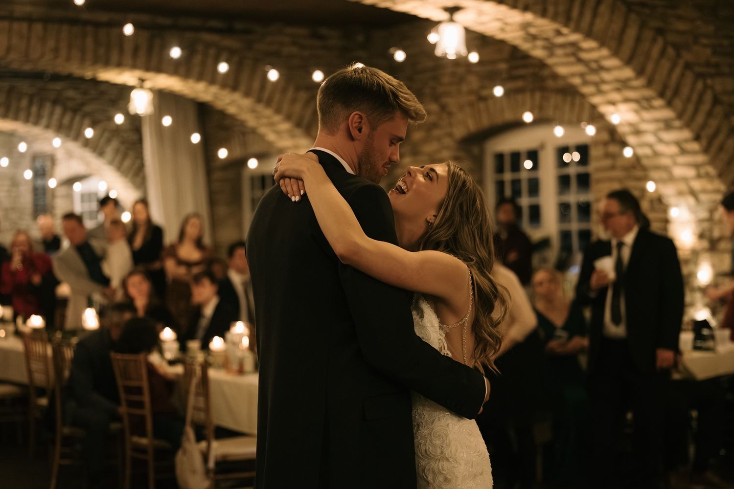 Couple shares first dance in romantic brick venue with string lights and wedding guests seated at tables.