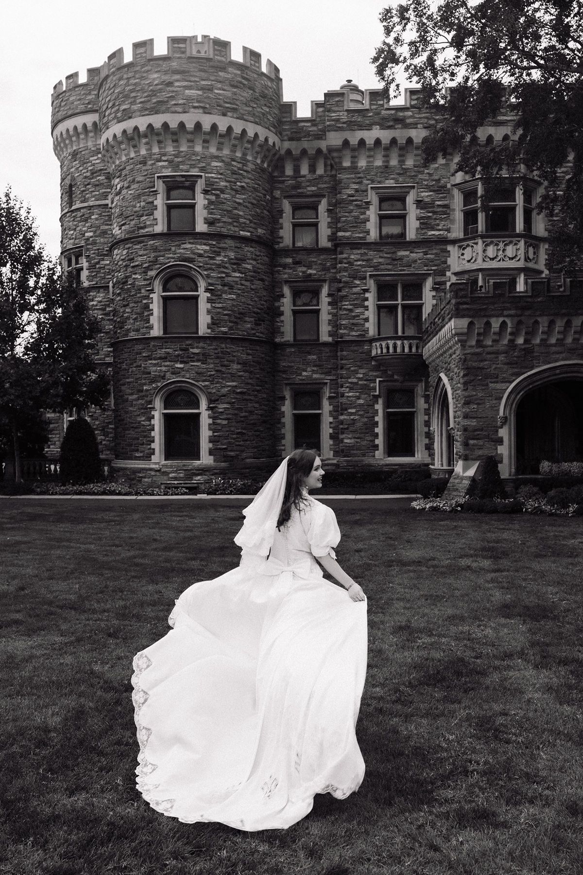Black and white photo of a bride in a flowing white gown and veil posing on a lawn before a grand stone castle.