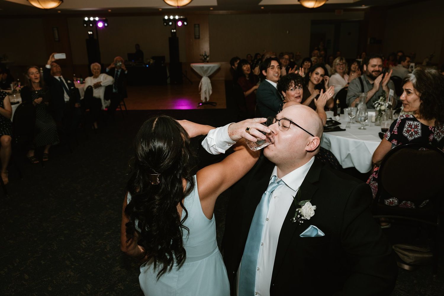 Wedding guests celebrate at a reception as a couple participate in a fun cake feeding moment on the dance floor.