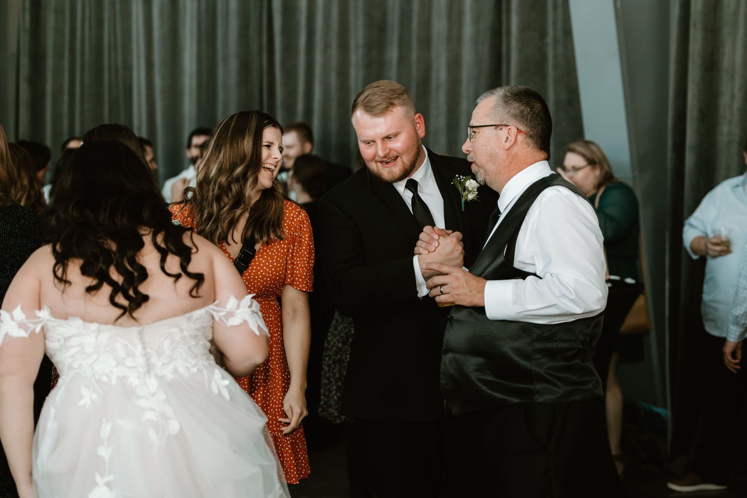 Wedding guests dance and mingle during a wedding reception celebration in a dimly lit venue with green curtains.