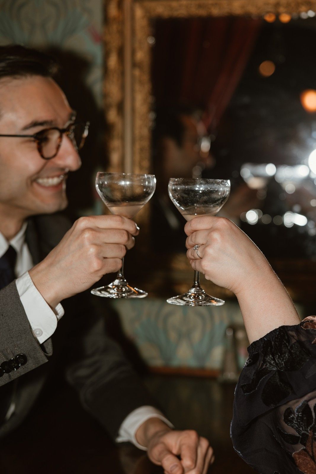 Two people toast with champagne coupes in a dimly lit vintage bar setting.