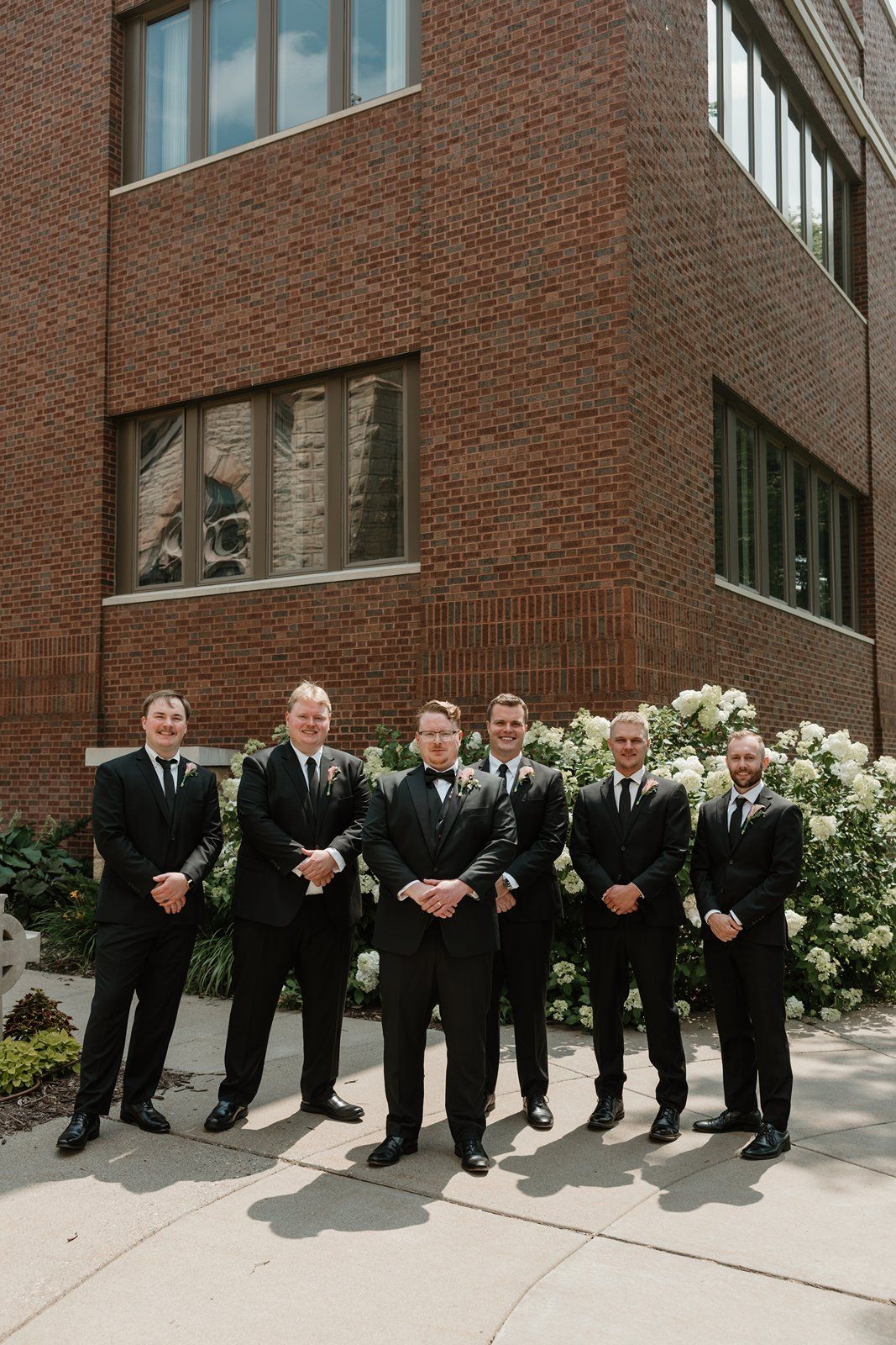 A group in black suits and ties stand in line against a brick wall with white flowers in a formal wedding party pose.