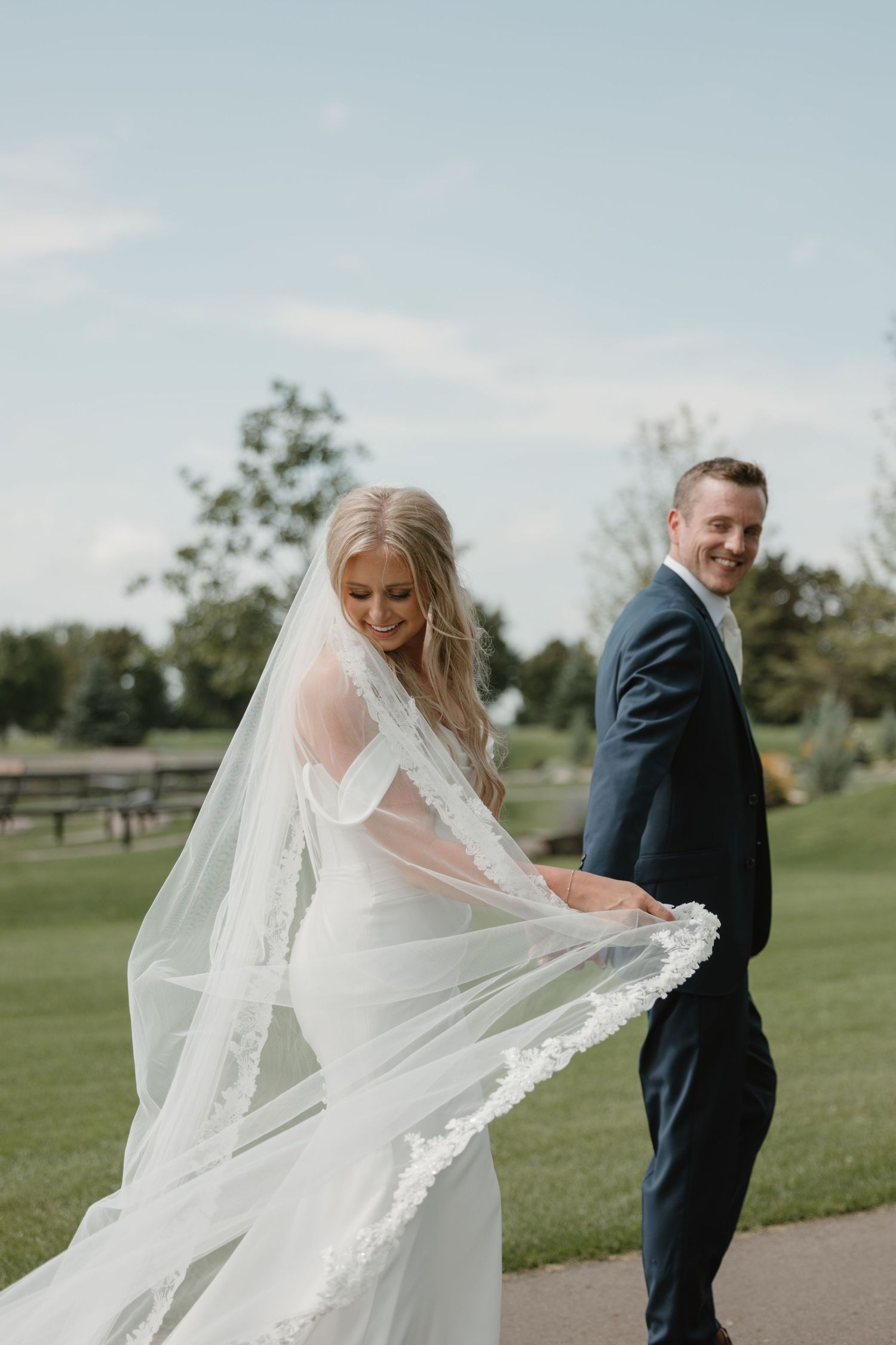 A couple on their wedding day walking outdoors on a path with a flowing lace veil catching the breeze.